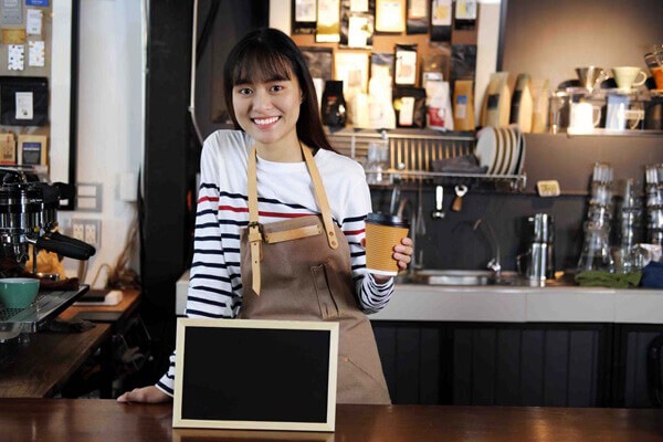 Portrait of smiling asian barista holding cup of coffee with blank chalkboard menu at counter in coffee shop. Cafe restaurant service, food and drink industry concept.